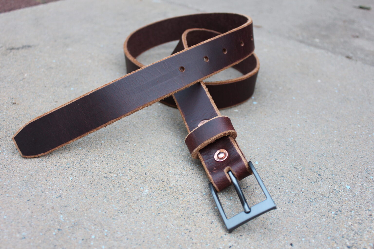 A single handmade leather belt coiled on concrete with the buckle facing forward
