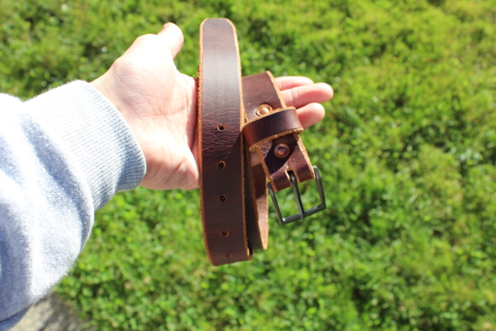A dark handmade full-grain leather belt held up against a green hedge