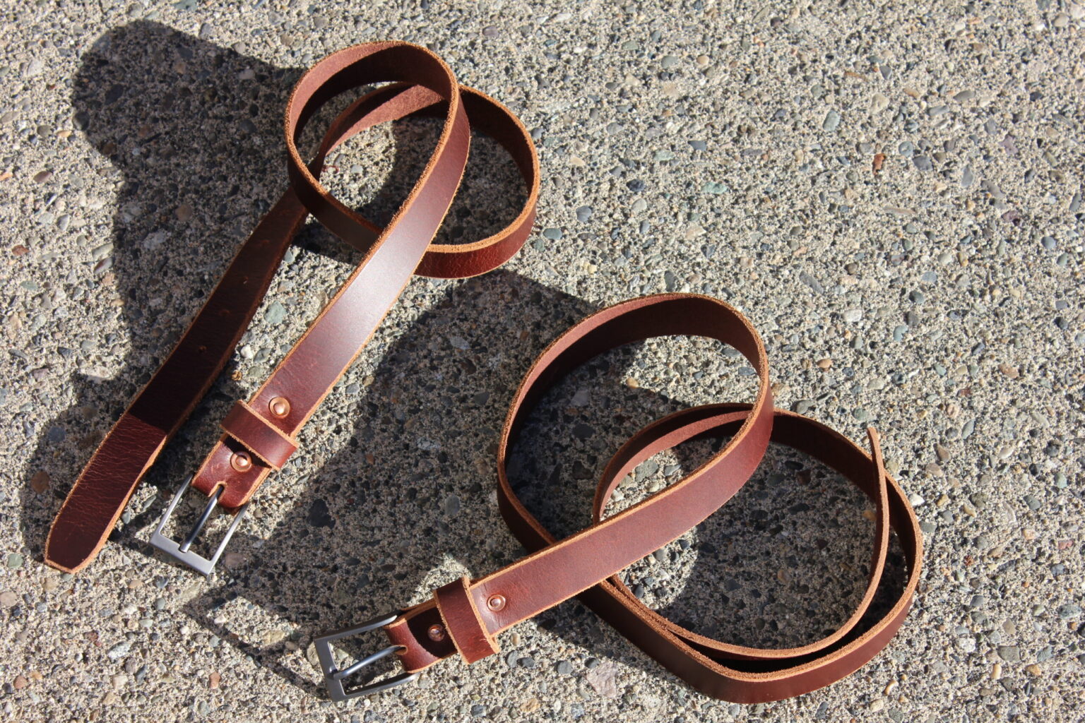 Two handmade leather belts laid out on sunlit asphalt, throwing long shadows